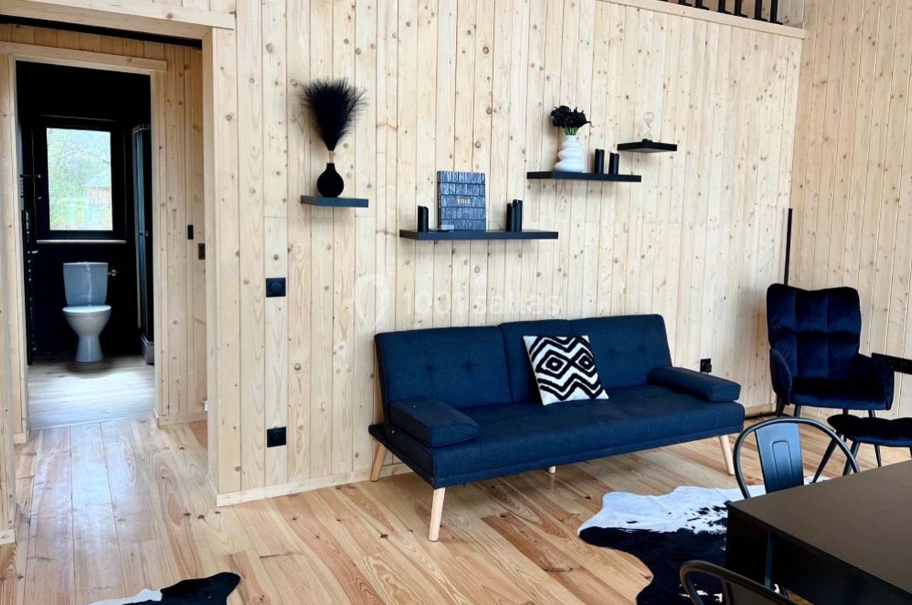 Salon en bois clair avec canapé bleu, étagères murales noires, table, chaises et vue sur une salle de bain au fond.