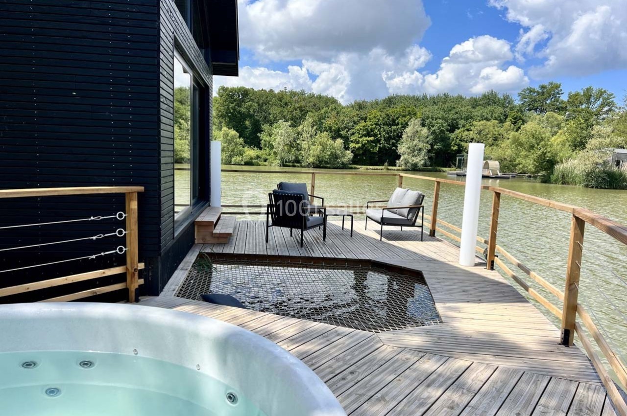 Terrasse en bois avec jacuzzi, fauteuils et filet suspendu, donnant sur un lac entouré de végétation.