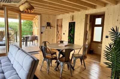 Intérieur d'une cabane en bois avec table ronde, chaises métalliques, canapé et terrasse extérieure visible.