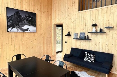 Intérieur d'une cabane en bois avec table ronde, chaises métalliques, canapé et terrasse extérieure visible.