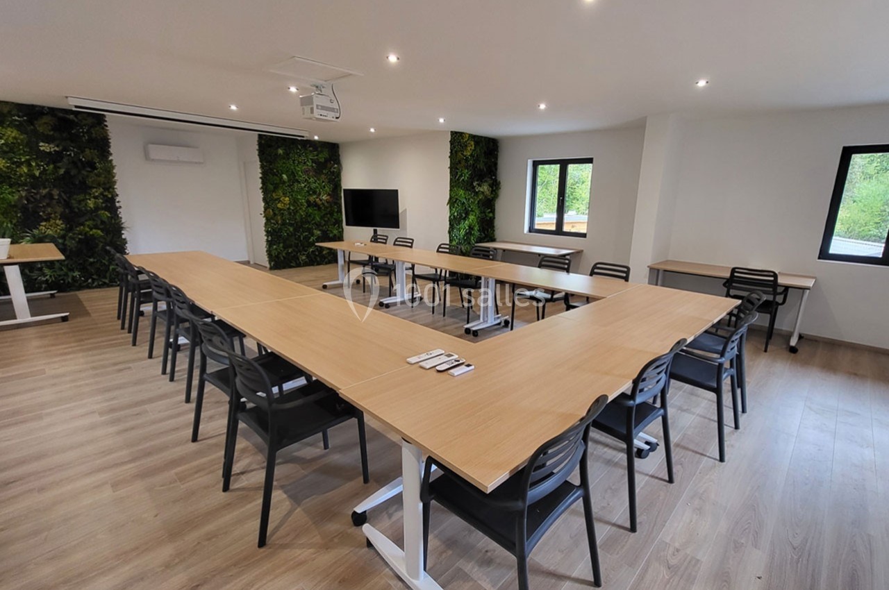 Salle de réunion lumineuse avec tables disposées en U, chaises noires, écran et murs végétalisés.