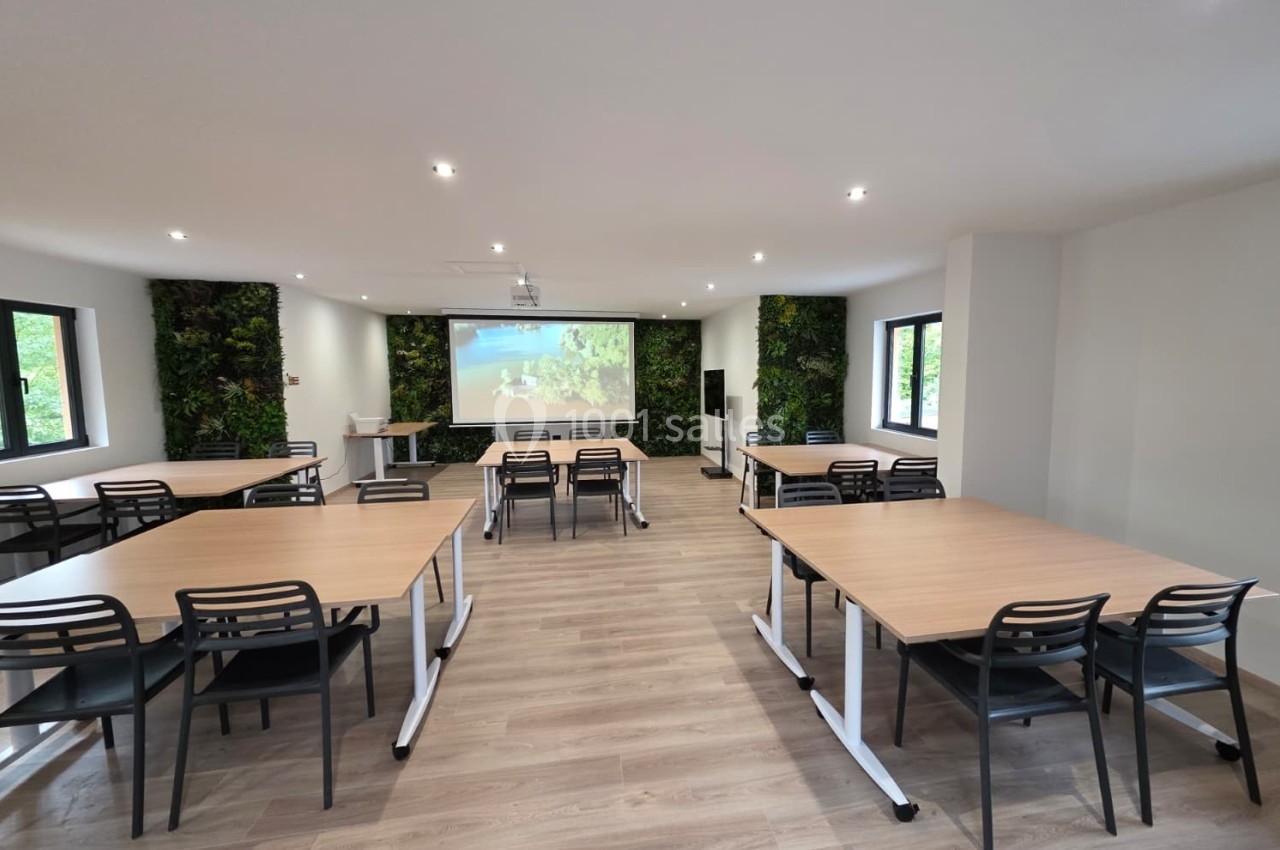 Salle de réunion lumineuse avec tables en bois, chaises noires, mur végétal et écran de projection.
