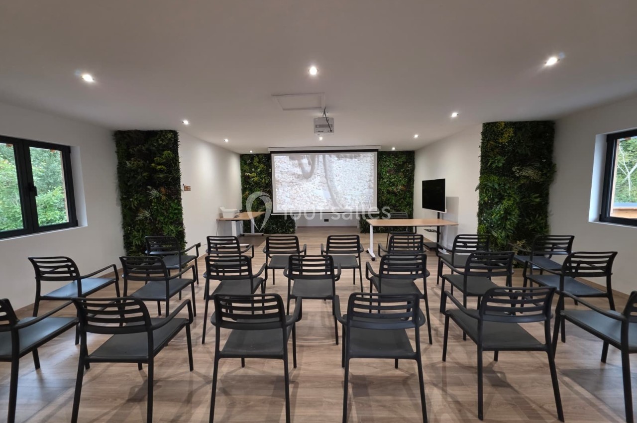 Salle de conférence lumineuse avec des rangées de chaises noires, un écran de projection et des murs végétalisés.