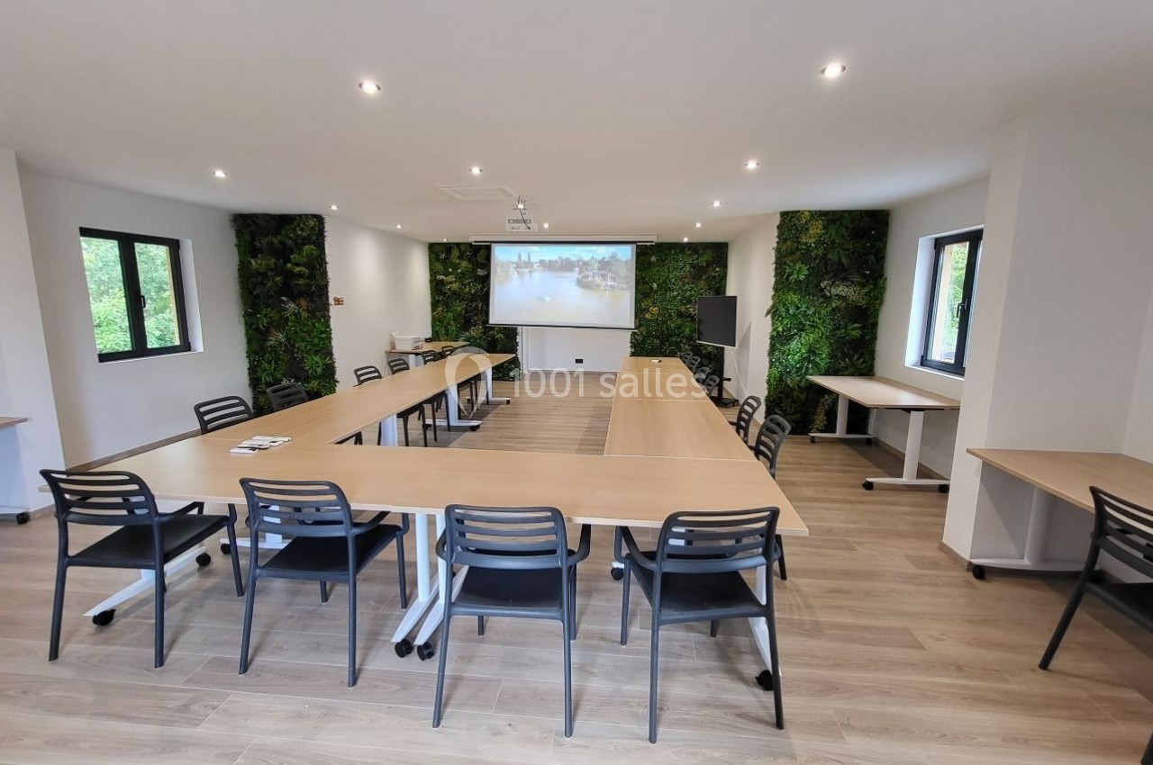 Salle de réunion lumineuse avec tables en U, chaises noires, mur végétal et écran de projection allumé.