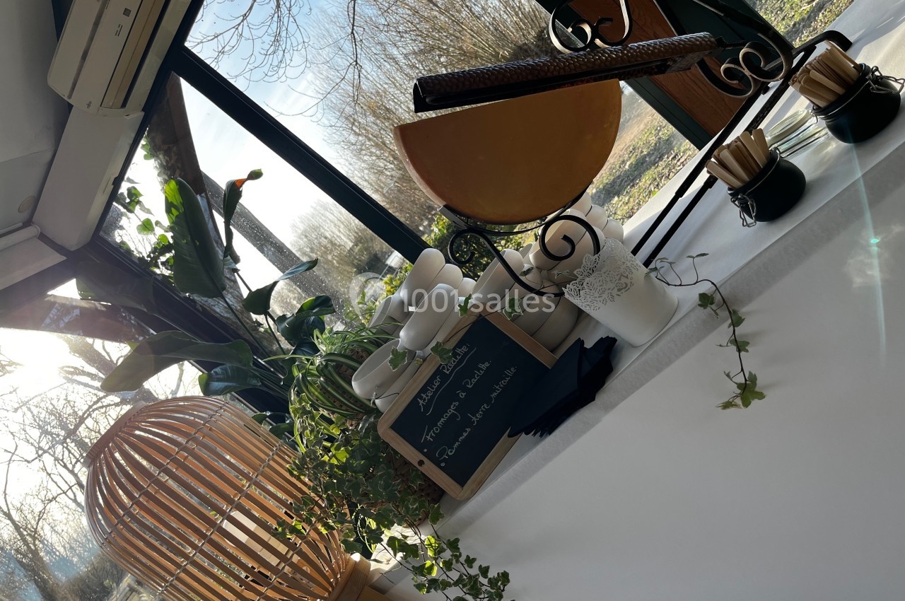 Table décorée avec plantes, vaisselle et une meule de fromage, près d'une baie vitrée donnant sur un paysage arboré.