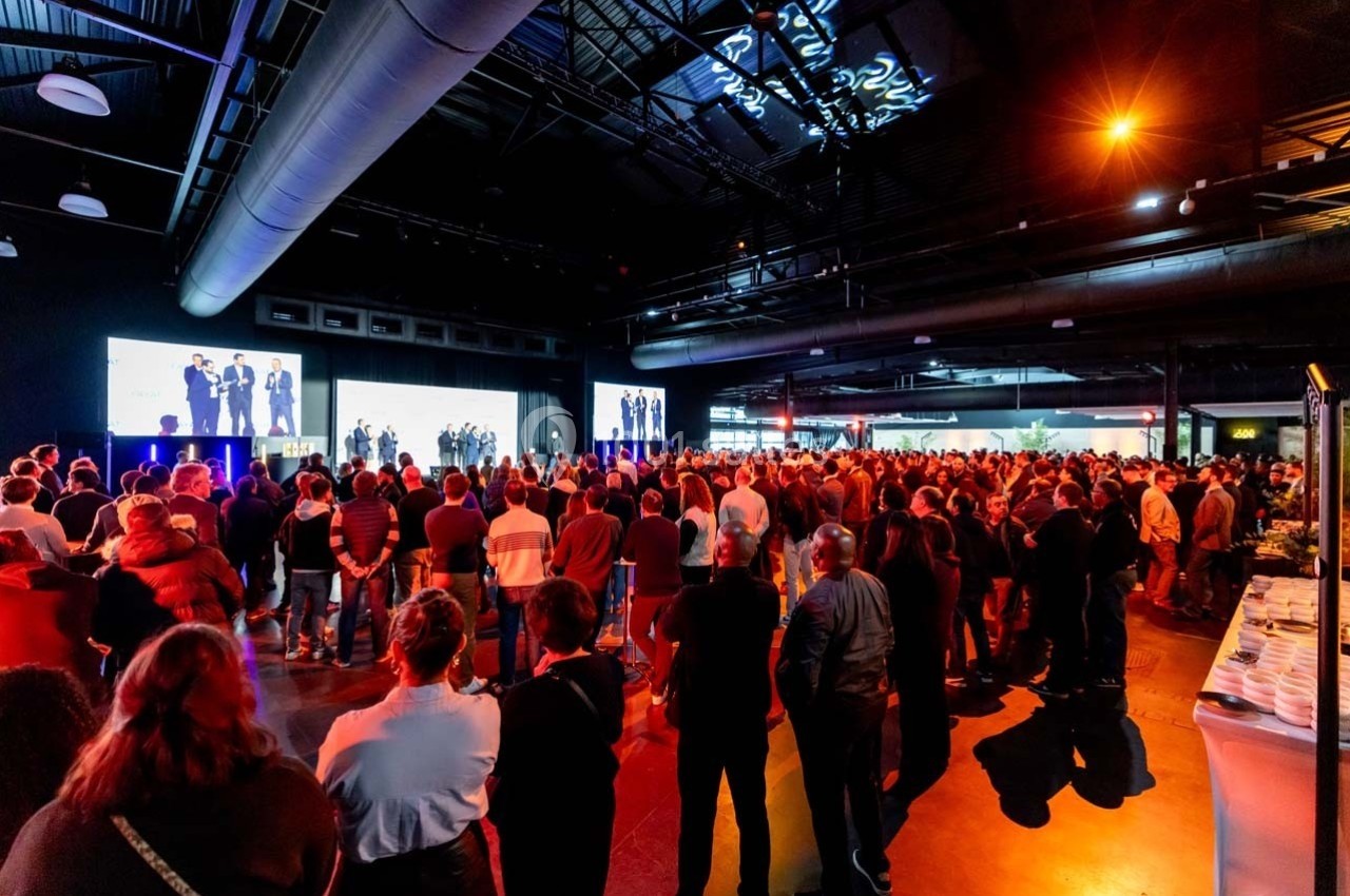 Un groupe de personnes assiste à une présentation sur scène dans une grande salle éclairée.