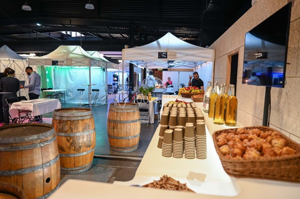 Buffet avec viennoiseries, bouteilles d'huile et gobelets alignés, dans un espace événementiel avec stands et tonneaux.