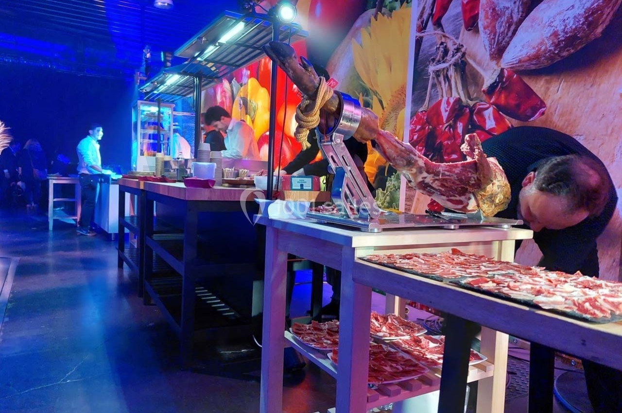 Découpe de jambon sur un stand de restauration dans un espace éclairé, avec des tables et des décorations colorées en…