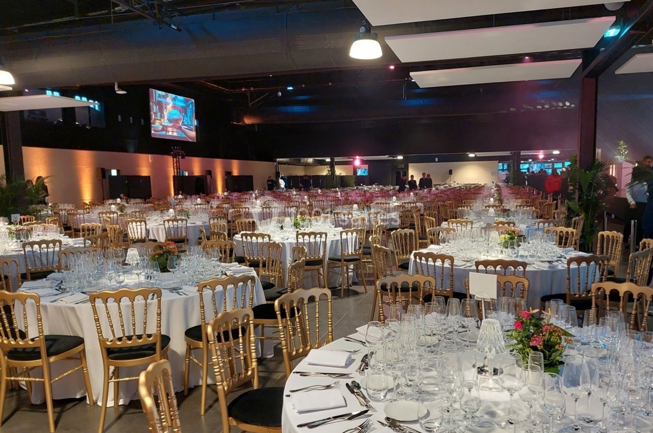 Salle de réception élégamment décorée avec des tables rondes dressées, chaises dorées et éclairage tamisé.