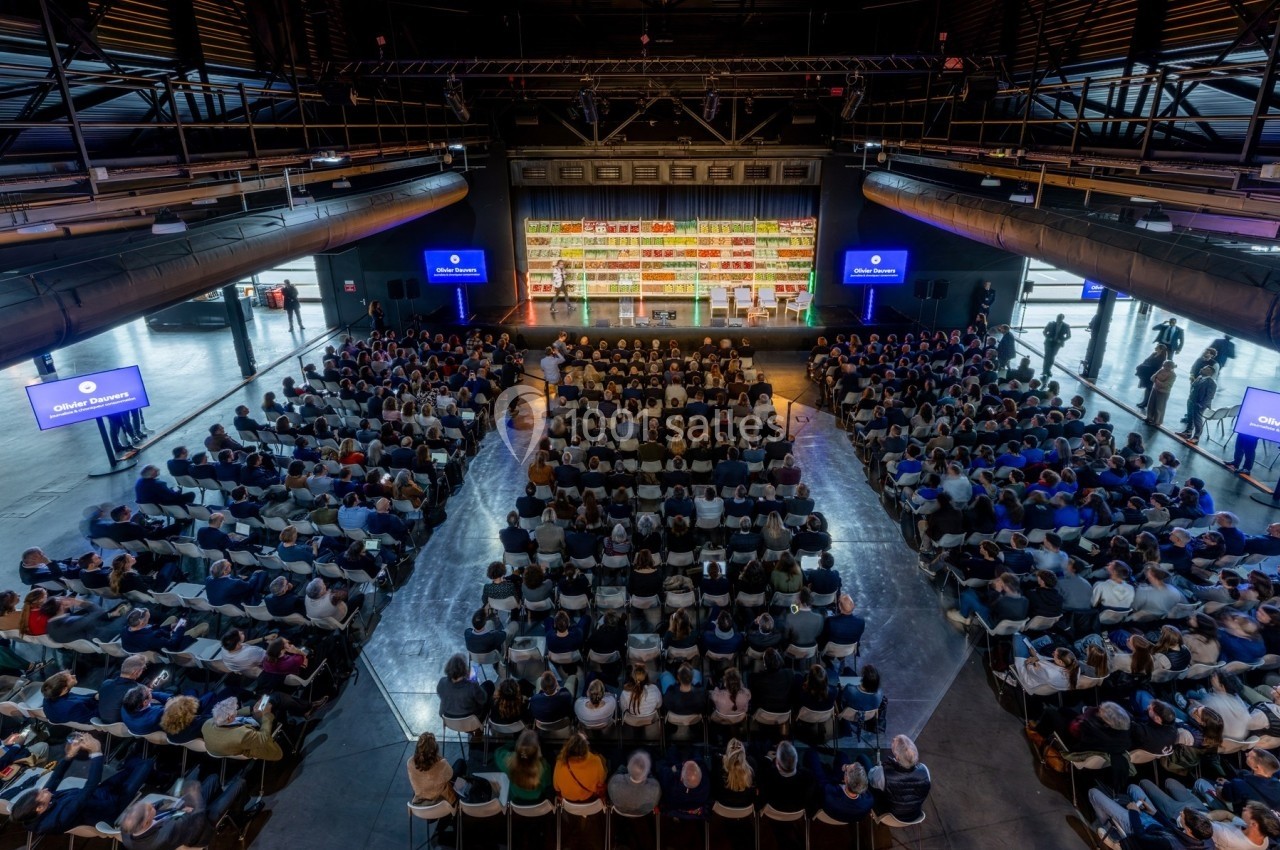 Salle comble avec un public assis face à une scène, entourée d'écrans et d'étagères colorées en arrière-plan.
