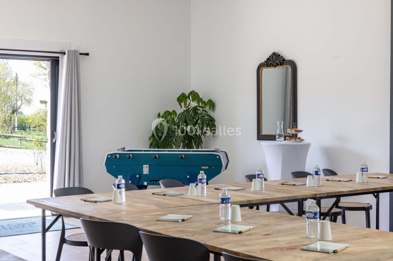 Salle lumineuse aménagée avec des tables en bois, des chaises, un baby-foot, un miroir et des bouteilles d'eau.