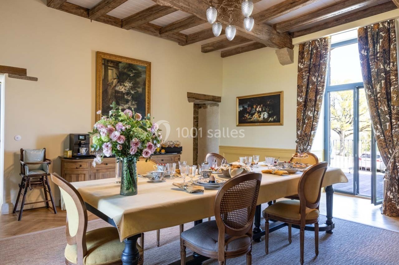 Salle à manger lumineuse avec une grande table dressée, bouquet de fleurs au centre, poutres apparentes et vue sur l…