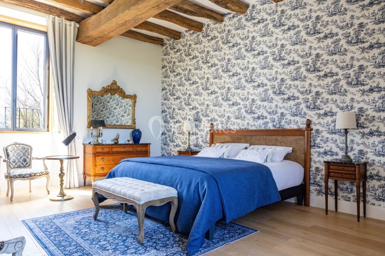 Chambre lumineuse avec lit double, couvre-lit bleu, mobilier en bois, mur tapissé et poutres apparentes.