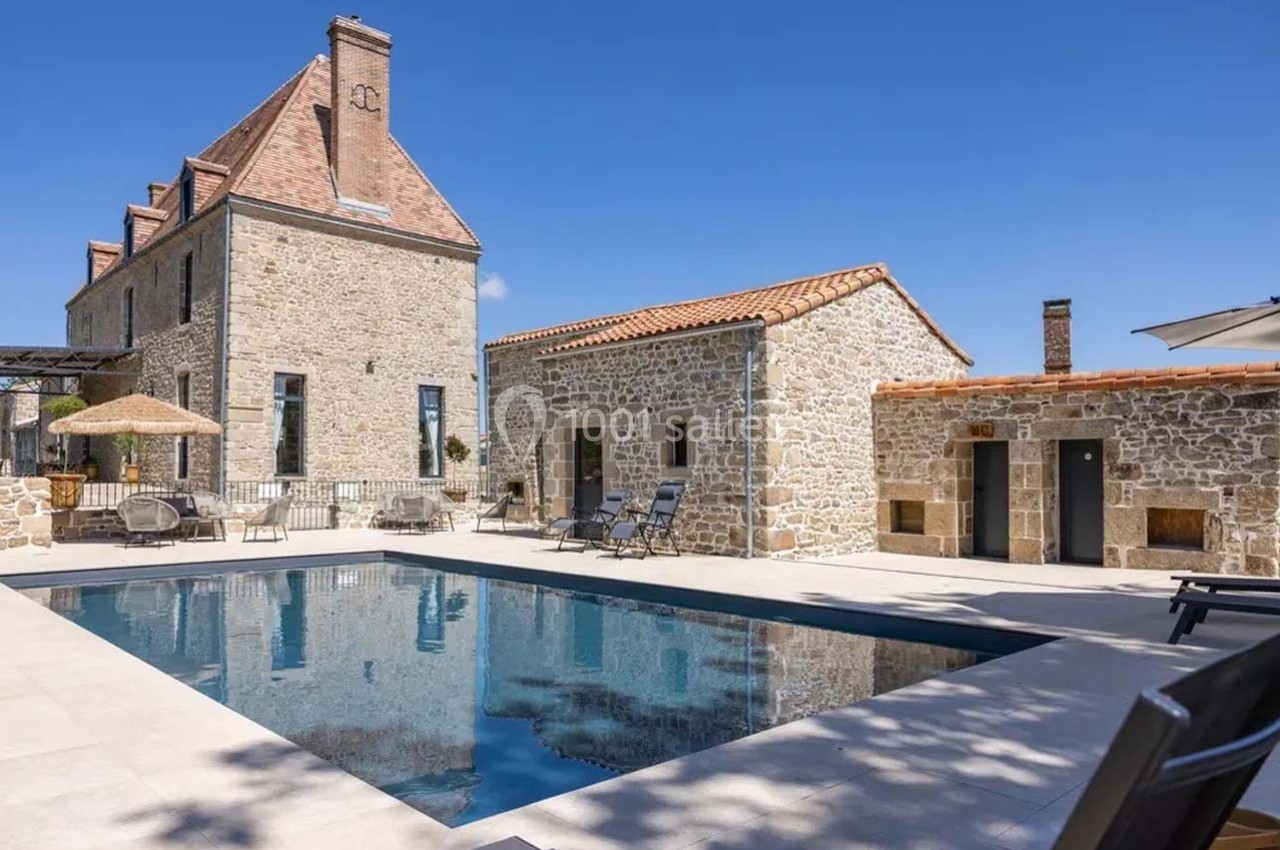 Piscine extérieure bordée de chaises longues devant une maison en pierre avec toit en tuiles sous un ciel bleu.
