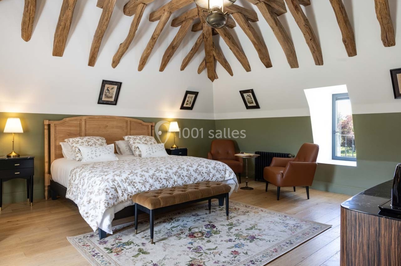 Chambre lumineuse avec poutres apparentes, lit double, fauteuils en cuir, tapis floral et fenêtres donnant sur l'extérieur.