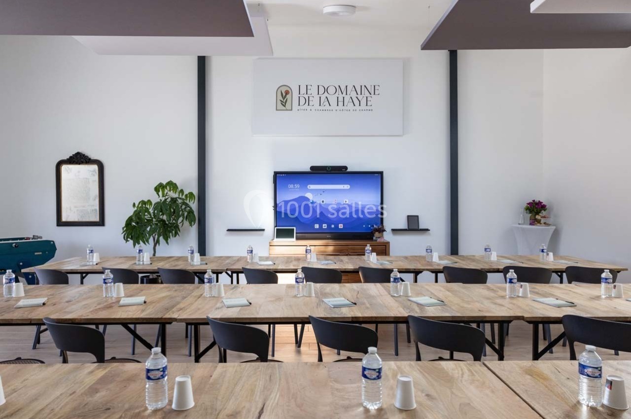 Salle de réunion lumineuse avec tables en bois, chaises noires, écran interactif et bouteilles d'eau disposées.
