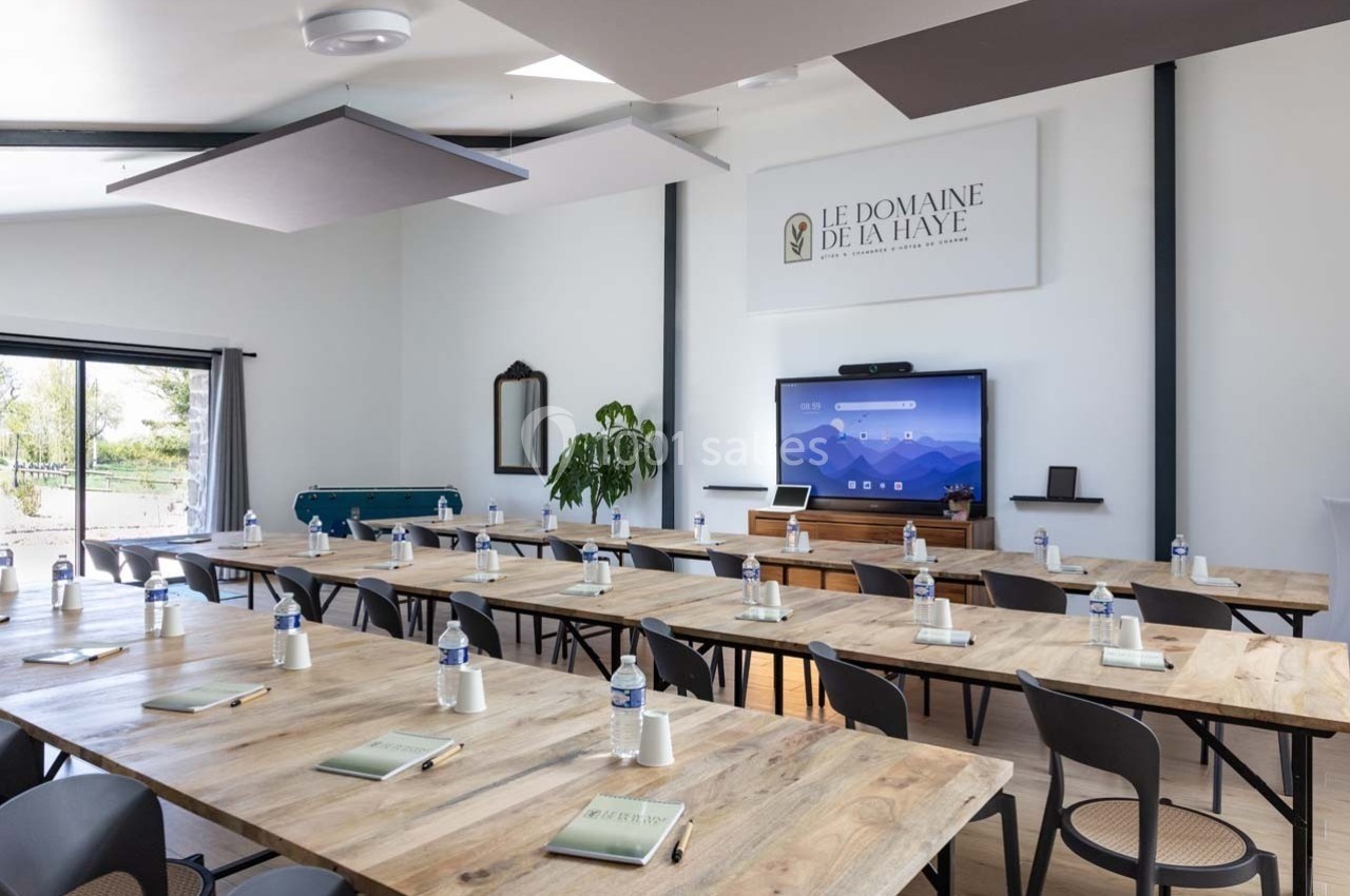 Salle de réunion lumineuse avec tables en bois, chaises noires, écran interactif et matériel de conférence disposé.