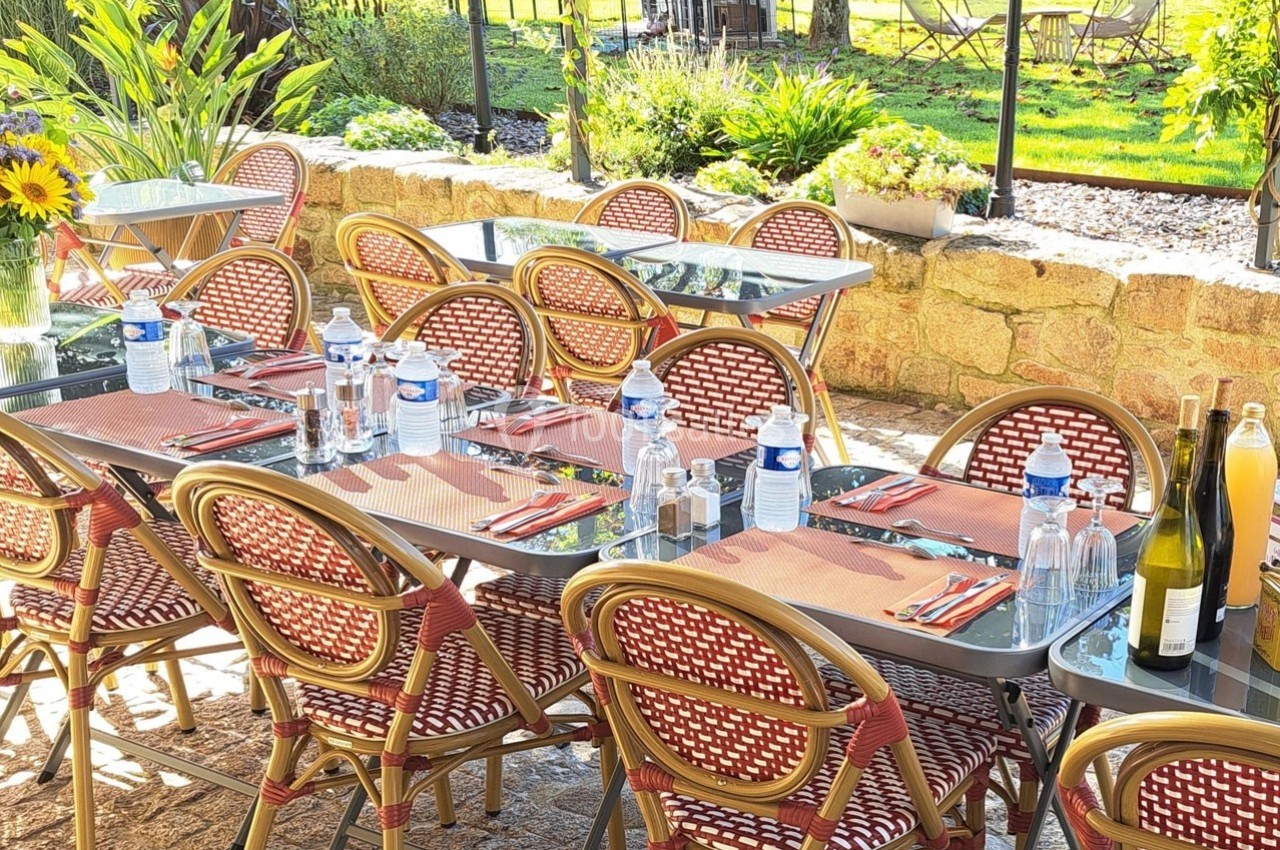 Terrasse ensoleillée avec tables dressées, chaises en osier, bouteilles d'eau et de vin, vue sur un jardin verdoyant.