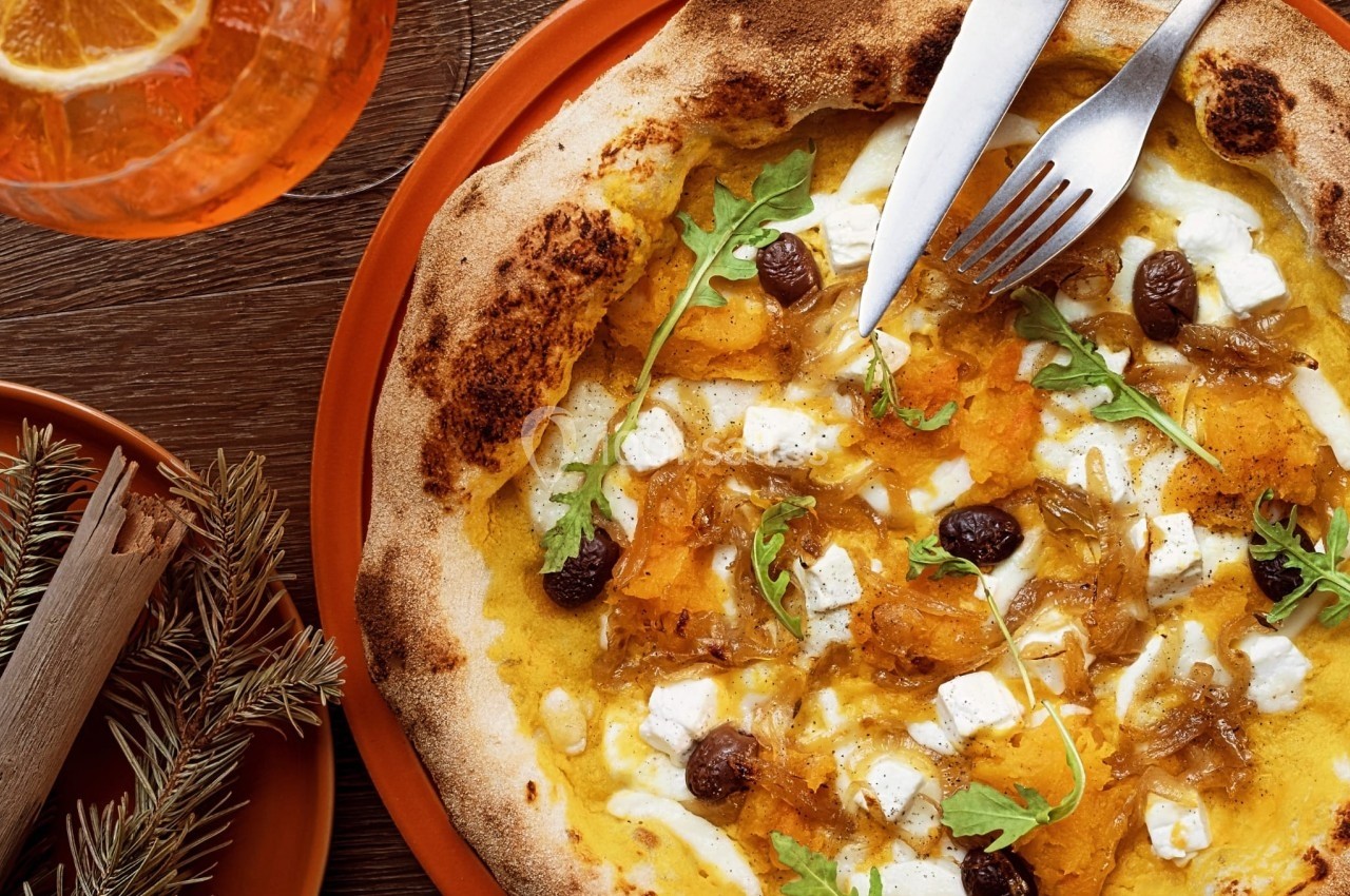 Pizza garnie de fromage, roquette, olives et morceaux de courge, servie avec un verre de boisson orange.