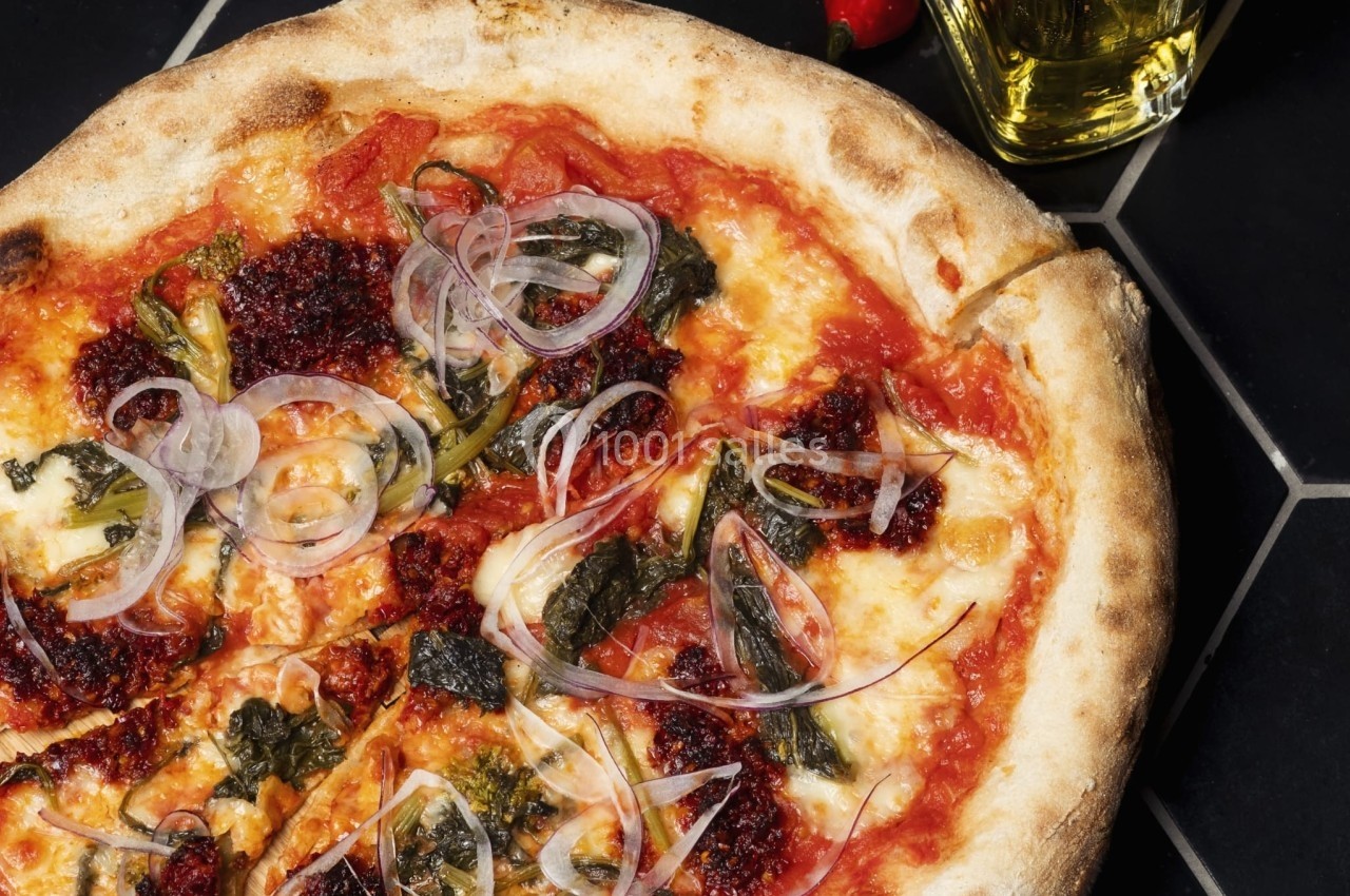 Pizza garnie de sauce tomate, fromage, oignons rouges, légumes verts et morceaux de piment, sur une table noire.