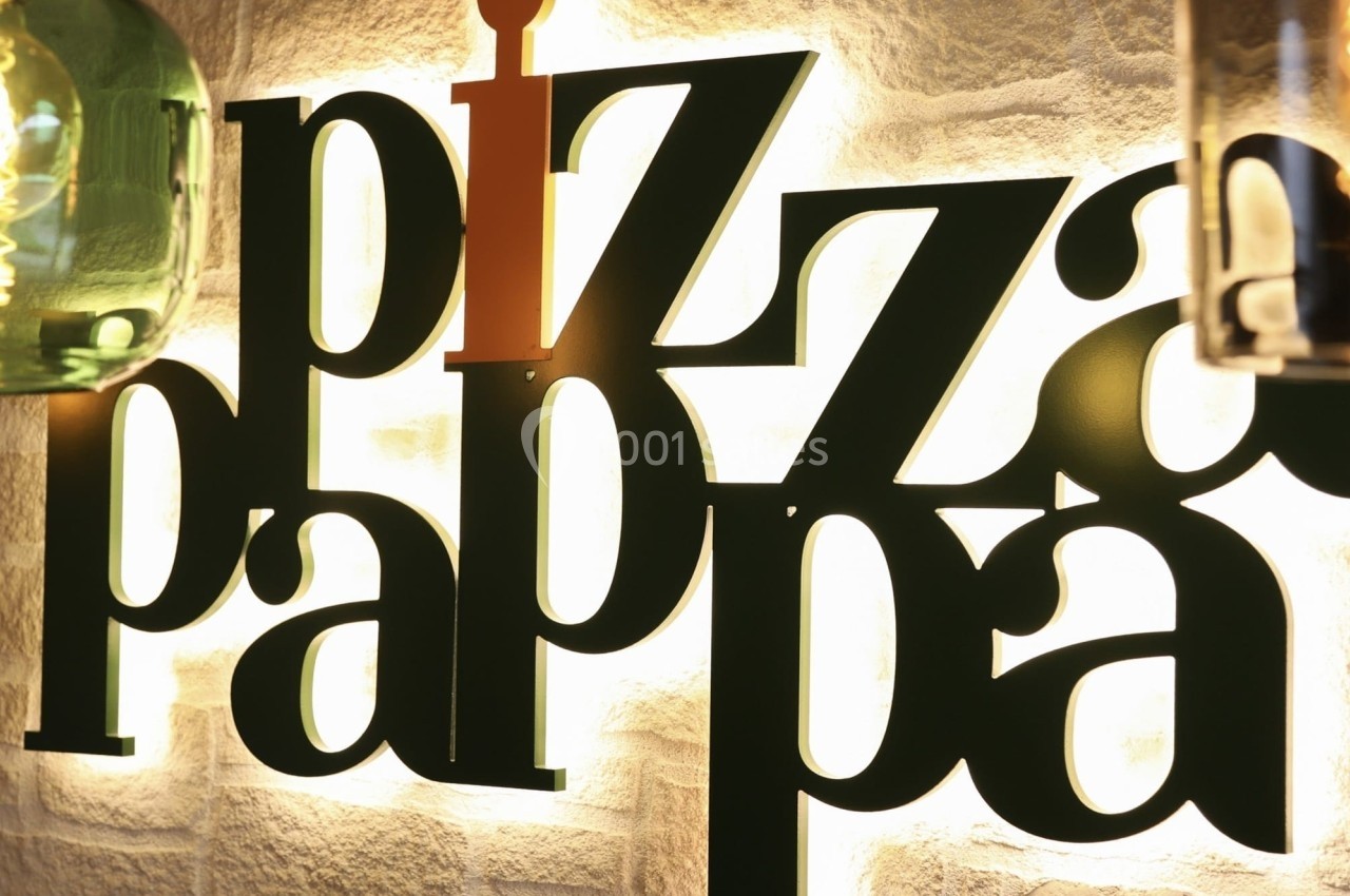 Enseigne lumineuse ’pizza pappa’ fixée sur un mur en pierre, éclairée par des lampes suspendues.