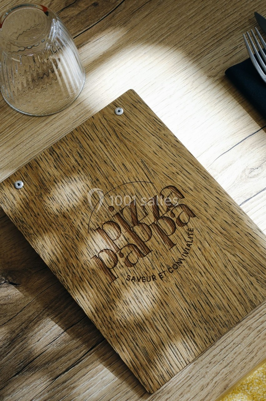 Menu en bois gravé ’Pizza Papa’ posé sur une table en bois clair, entouré de couverts et d'un verre retourné.
