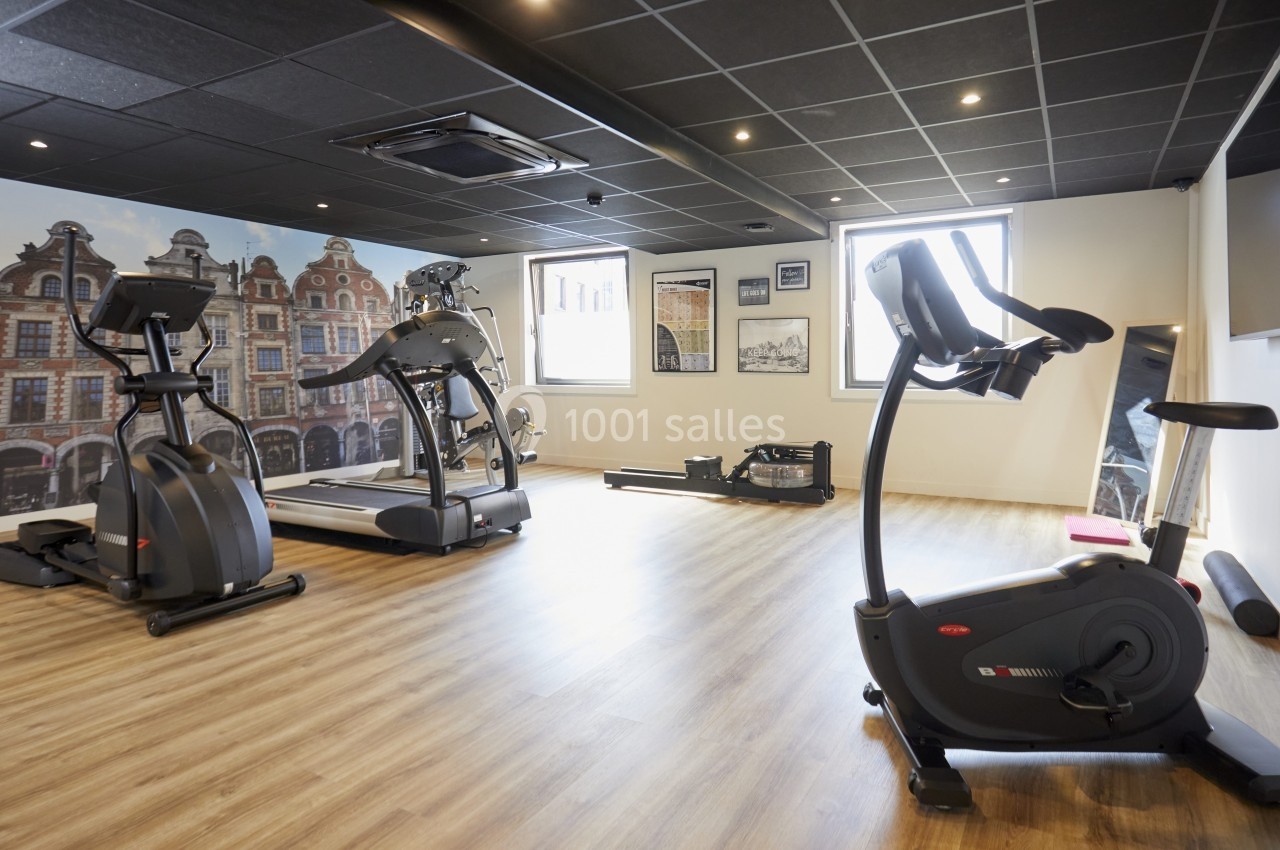Salle de sport lumineuse avec tapis de course, vélos d'exercice et rameur sur un sol en parquet.