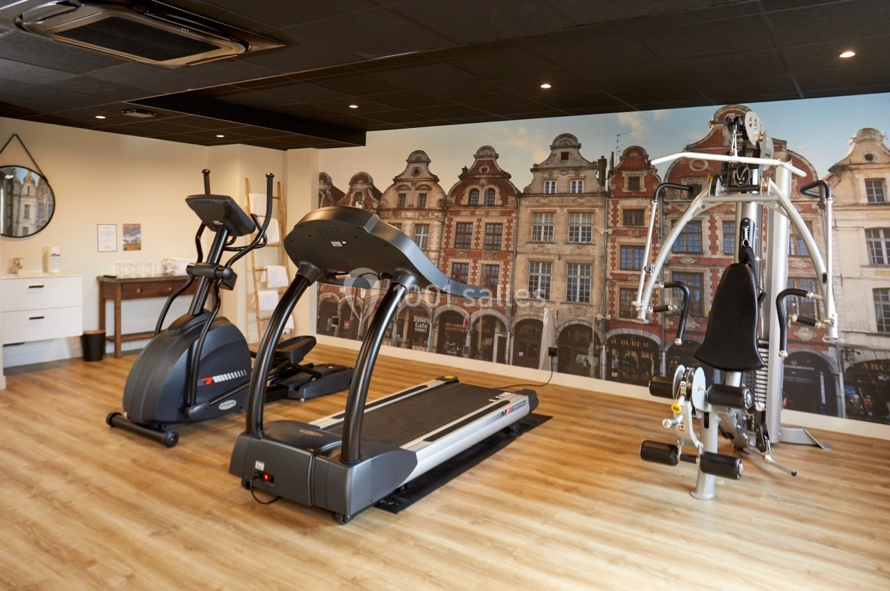 Salle de sport équipée d'un tapis de course, d'un vélo elliptique et d'une machine de musculation, avec décor mural urbain.