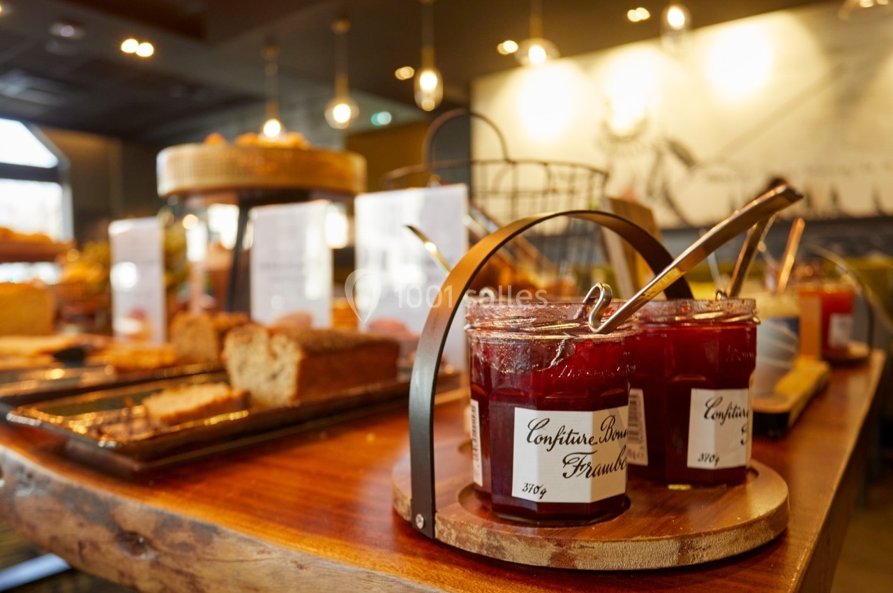 Pots de confiture de fraise avec cuillères sur une table en bois, entourés de pains et viennoiseries dans un buffet.