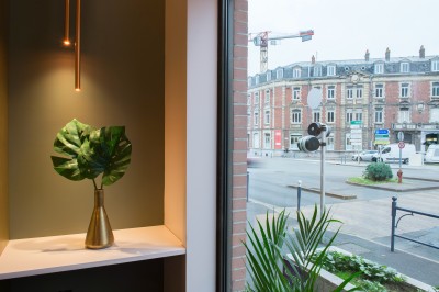 Feuilles vertes dans un vase doré sur une étagère près d'une fenêtre donnant sur une rue urbaine avec bâtiments et grue.