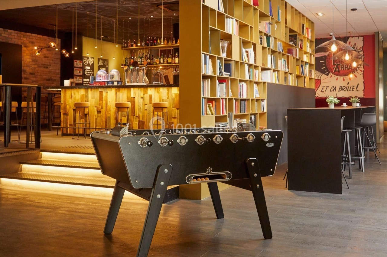 Baby-foot noir dans un espace convivial avec bar en bois, étagères remplies de livres et éclairage chaleureux.
