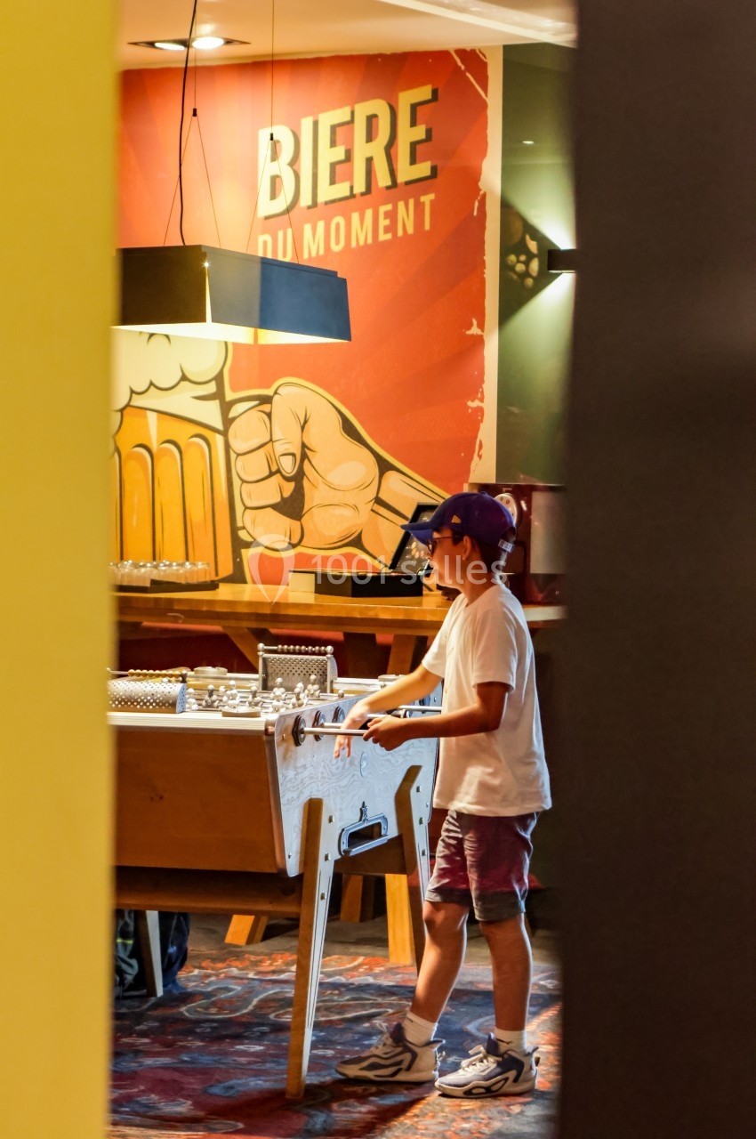 Un enfant joue au baby-foot dans un bar avec une affiche ’Bière du moment’ en arrière-plan.