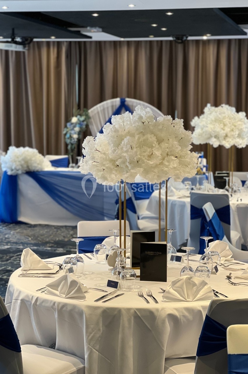 Salle de réception décorée pour un événement, avec tables dressées, nappes blanches et accents bleus, et centres de table…