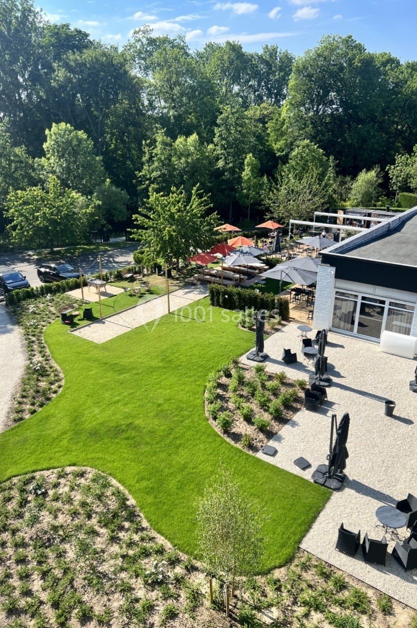 Vue aérienne d'une terrasse aménagée avec des tables, entourée de pelouses, d'arbustes et d'arbres en arrière-plan.