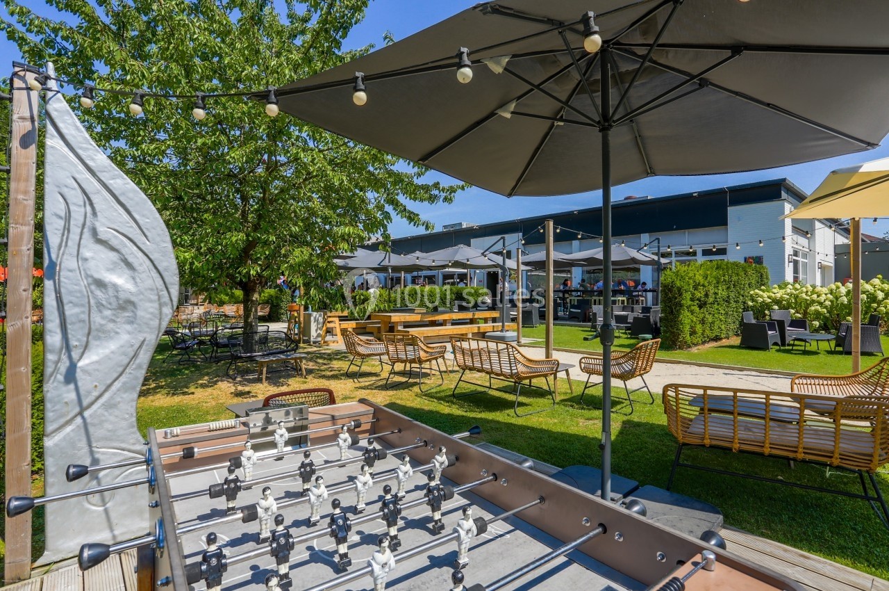 Terrasse extérieure avec baby-foot, parasols, mobilier de jardin et espace verdoyant sous un ciel ensoleillé.