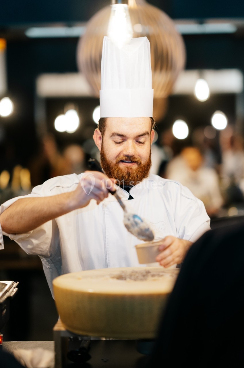 Un chef en tenue blanche sert un plat à partir d'une grande meule de fromage dans un environnement lumineux.