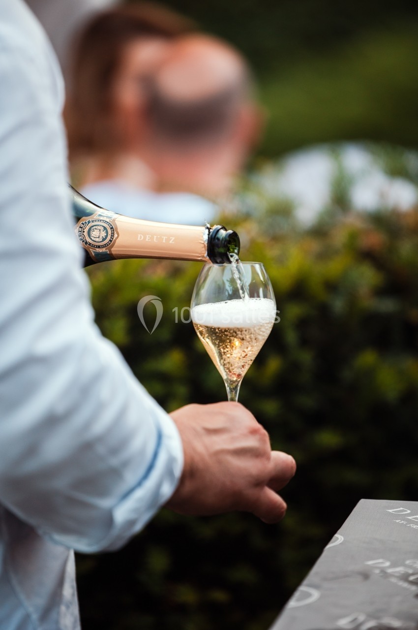 Une main verse du champagne pétillant dans une flûte, avec un arrière-plan flou de verdure et de personnes.