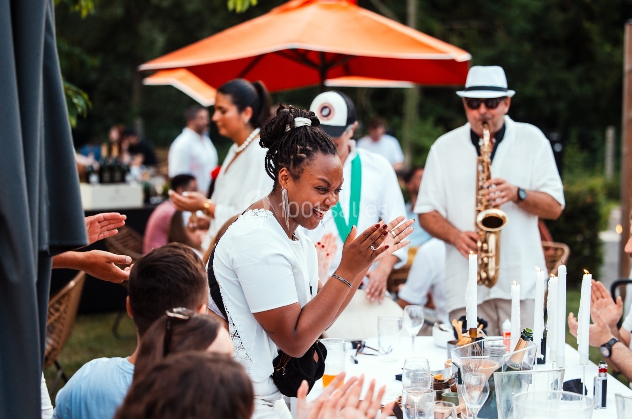Une femme souriante applaudit lors d'un repas en extérieur, avec un musicien jouant du saxophone en arrière-plan.