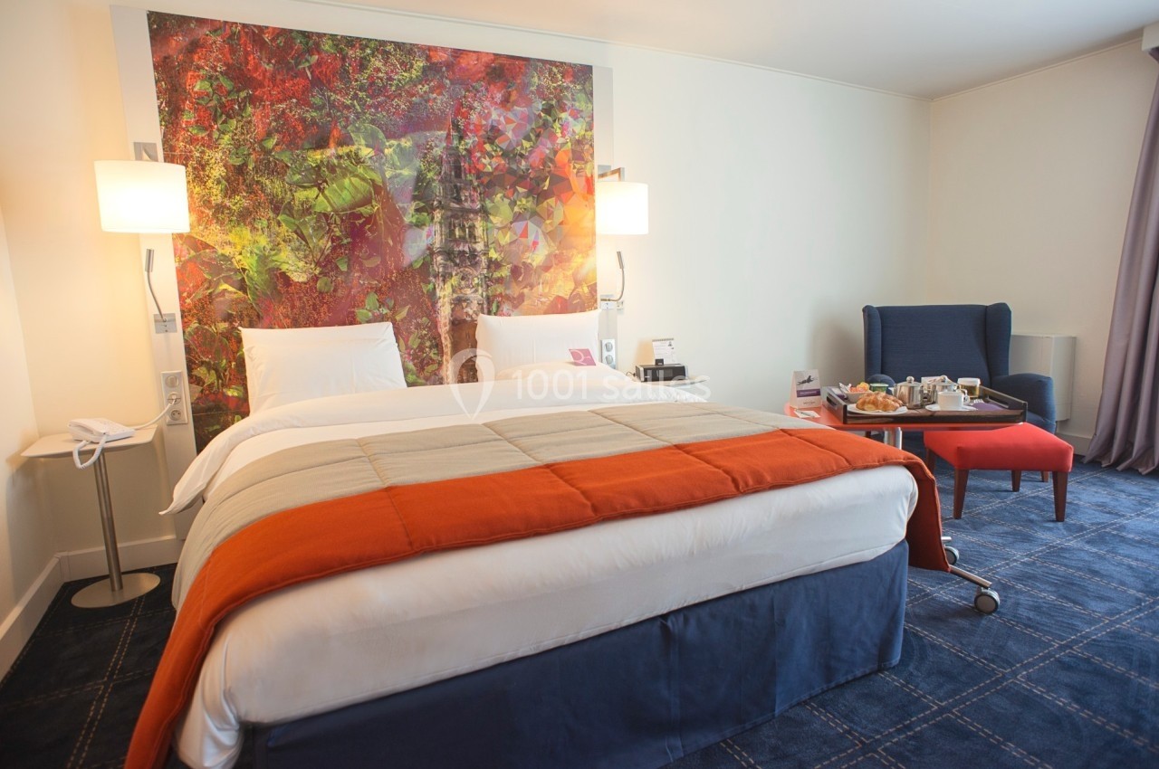 Lit double avec couvre-lit orange dans une chambre d'hôtel moderne, décorée avec un tableau coloré au mur.