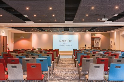 Salle de conférence avec des rangées de chaises colorées face à un écran de projection allumé.