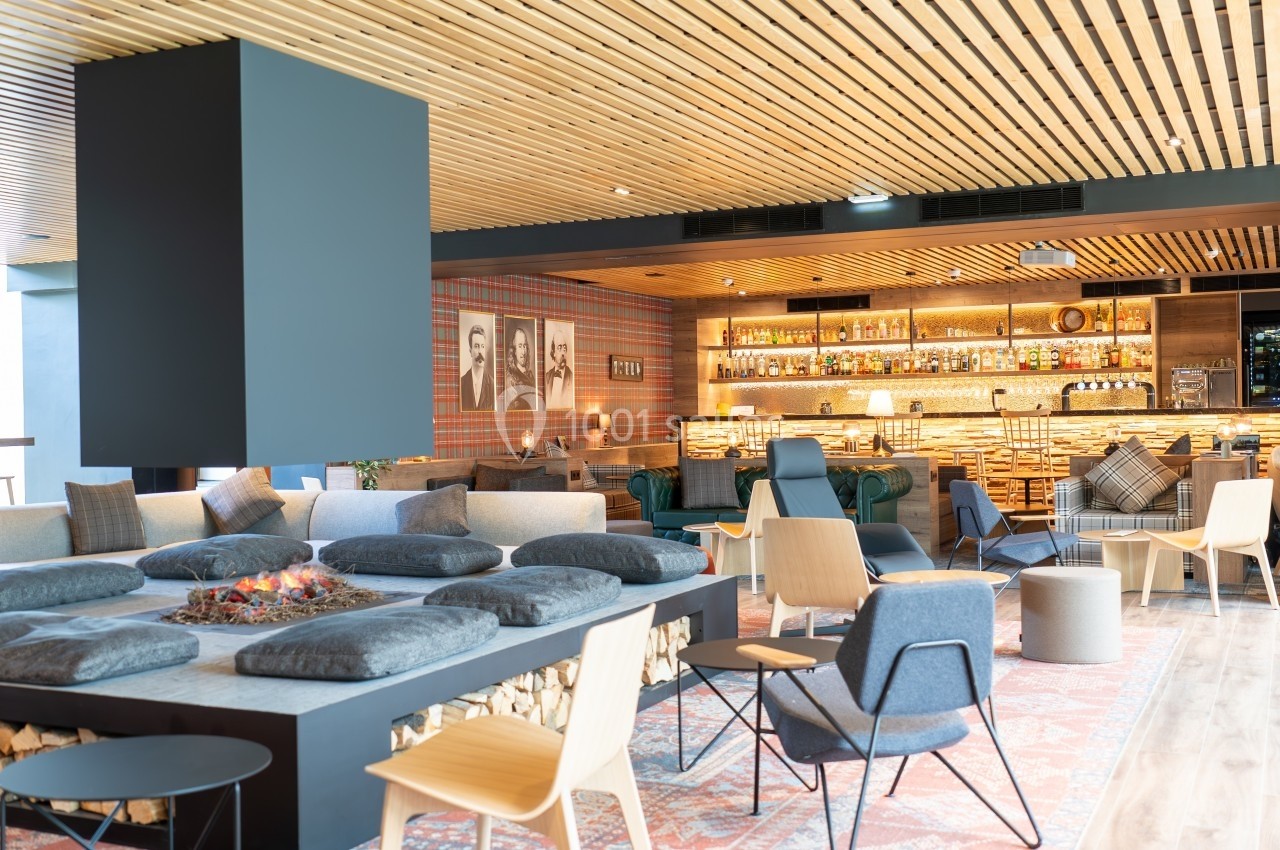 Espace lounge moderne avec cheminée centrale, fauteuils variés et bar en arrière-plan sous un plafond en bois.