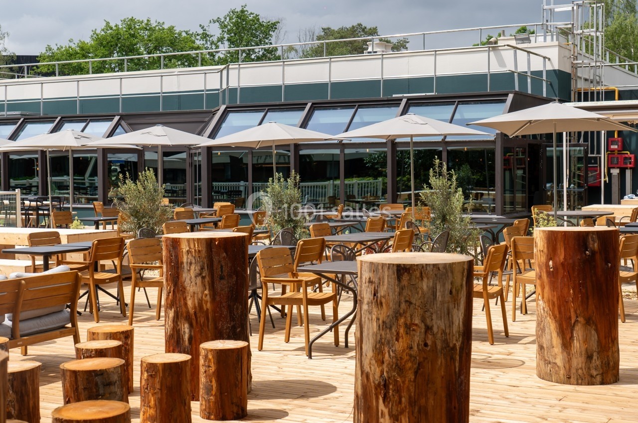 Terrasse en bois avec tables, chaises, parasols et troncs d'arbres décoratifs, entourée de verdure et d'un bâtiment moderne.