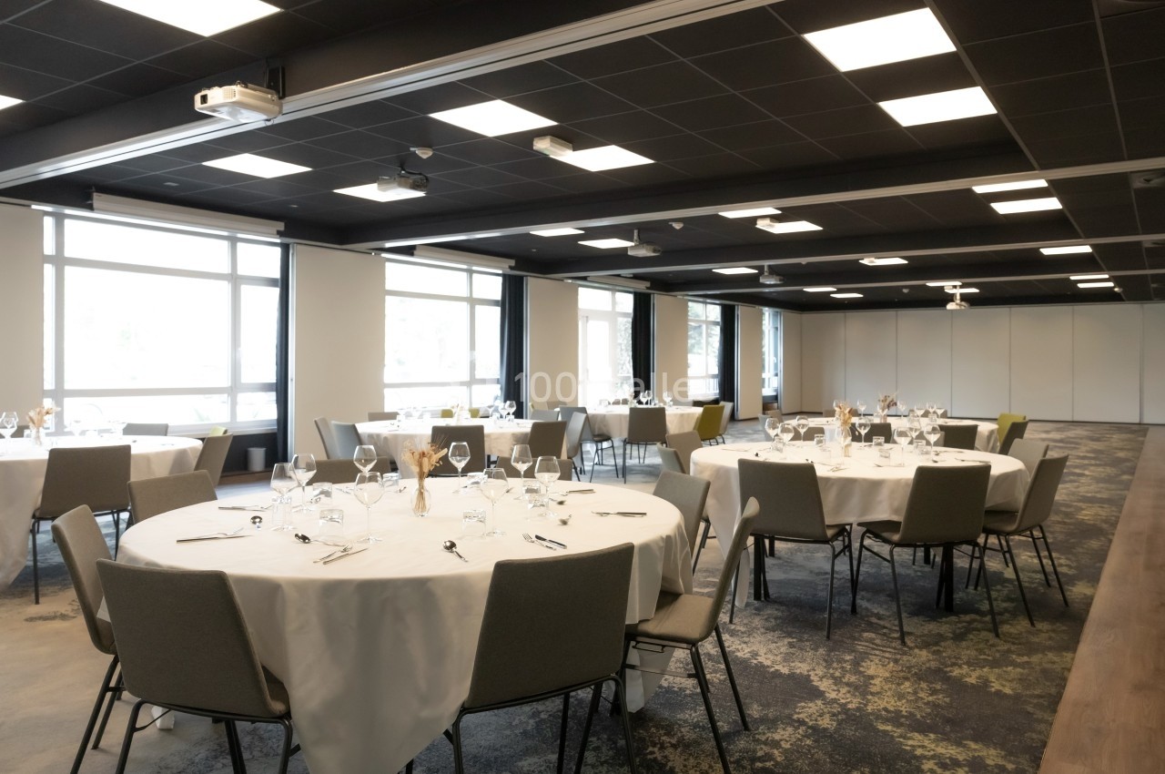 Salle de réception lumineuse avec tables rondes dressées, nappes blanches et chaises grises.