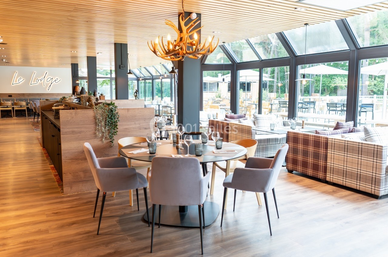 Salle de restaurant lumineuse avec grandes baies vitrées, mobilier moderne et lustre en forme de bois de cerf.