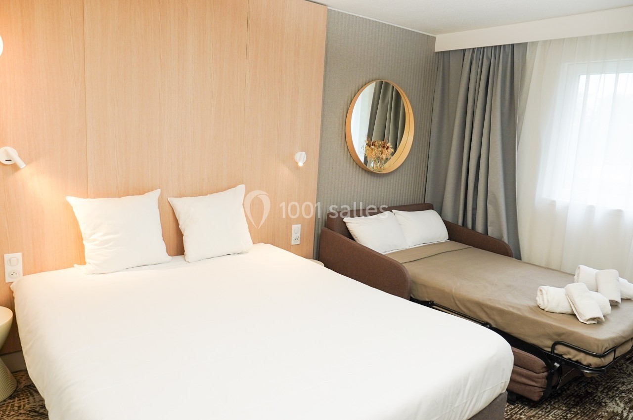 Chambre d'hôtel avec un lit double, un canapé-lit, un miroir rond et une fenêtre avec rideaux.