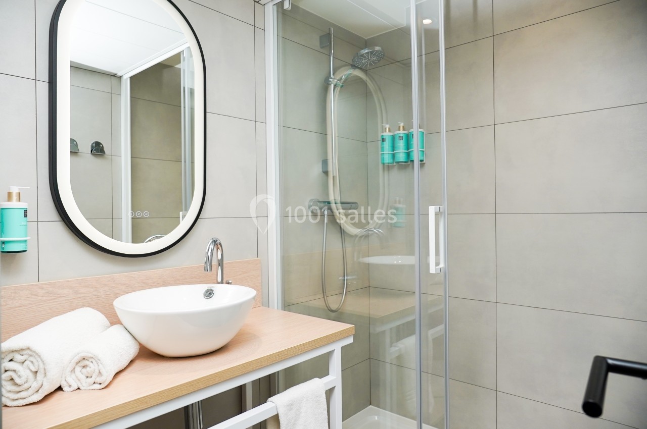 Salle de bain moderne avec lavabo, miroir ovale, douche vitrée et étagères murales contenant des produits.