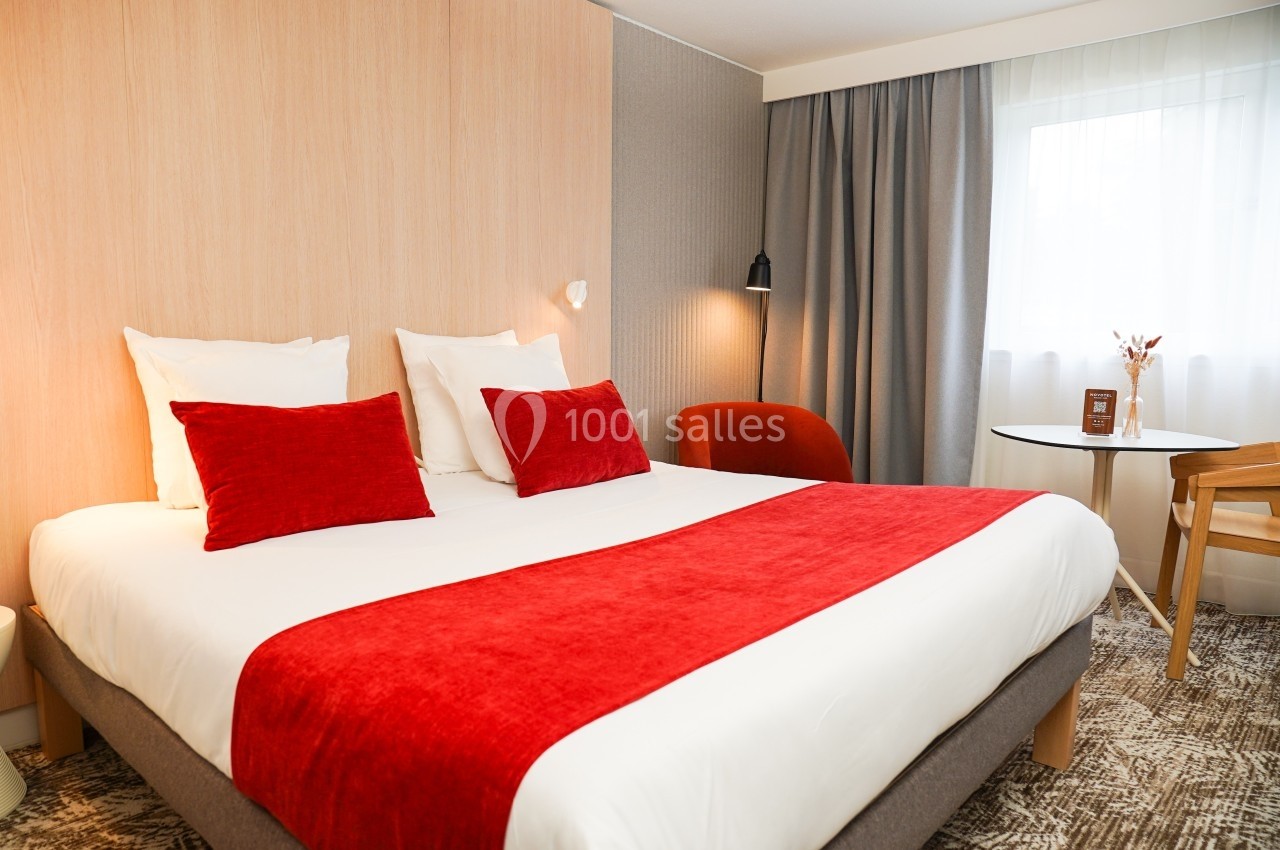 Chambre d'hôtel avec lit double, draps blancs, coussins et plaid rouges, table ronde, chaise et fenêtre avec rideaux.