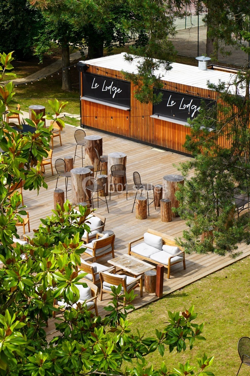 Terrasse en bois avec tables, chaises et fauteuils, entourée de verdure, devant un bâtiment marqué ’Le Lodge’.