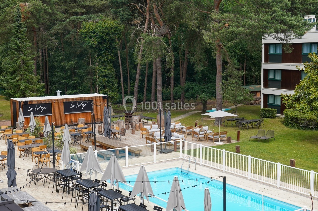 Terrasse avec piscine, chaises longues, tables et parasols, entourée de verdure et d'un bâtiment moderne.