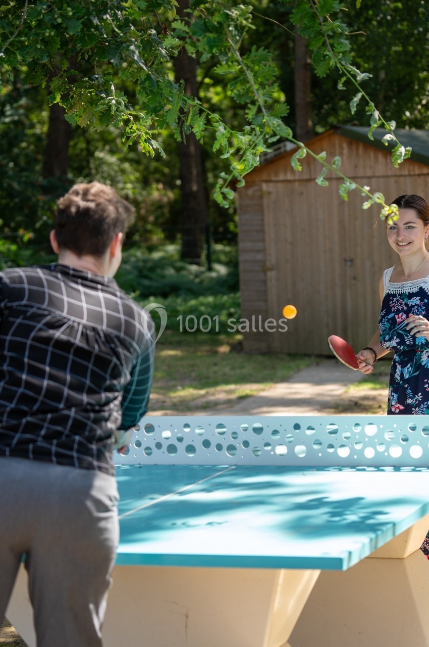 Deux personnes jouent au tennis de table en extérieur, près d'un abri en bois et entourées de verdure.